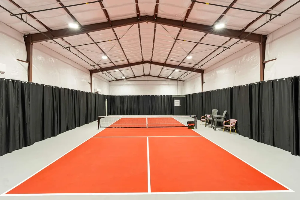 The private indoor pickleball court under bright lights, with an orange playing surface and black curtains along the walls